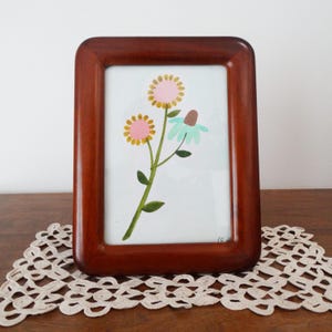 May include: A rectangular dark brown wooden picture frame containing a white print of a green stem with two yellow-petaled flowers with pink centers and one light blue flower with a brown center. The frame has rounded corners and a smooth finish.