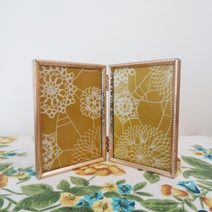 May include: A double photo frame with a gold-coloured metal frame, hinged in the middle. The frame contains two mustard-yellow panels with a white floral pattern. The frame is open, displaying the patterned fabric.