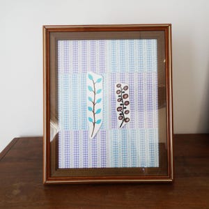 May include: A rectangular wooden frame containing a collage with a background of vertical light blue and purple checkered stripes. Two paper cutouts are centered: one tall branch with light blue leaves and one shorter branch with brown circles.