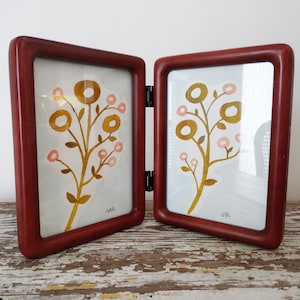 May include: A hinged, double-sided picture frame with a dark red wooden finish. Each side displays a watercolor painting of a floral design with gold and pink accents. The frame is open, revealing the artwork.