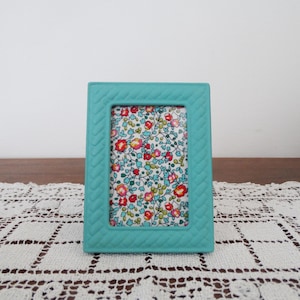May include: A turquoise picture frame with a textured border, holding a floral fabric print. The fabric features red, pink, and yellow flowers on a light blue backdrop. The frame is displayed on a white lace doily.