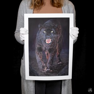 May include: A framed print depicting a black panther with yellow eyes and an open mouth, revealing its teeth. The artwork uses shades of black, brown, and red against a dark backdrop. The print is held by a person wearing a grey cardigan and white gloves.