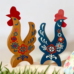 Könnte beinhalten: Zwei hölzerne Hahnfiguren. Eine ist aus Naturholz mit roten, blauen und weißen Blumenmustern. Die andere ist marineblau mit weißen und roten Blumenmustern. Beide haben rote Kämme und stehen auf einer hellen Oberfläche.
