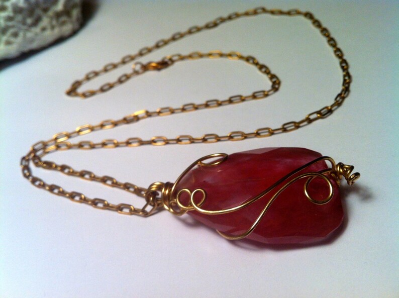 May include: A pink gemstone pendant necklace with a gold chain and wire wrapping. The gemstone is a unique shape and has a natural, organic look.