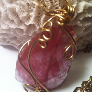 May include: A pink and white gemstone pendant wrapped in gold wire. The pendant is hanging from a gold chain.