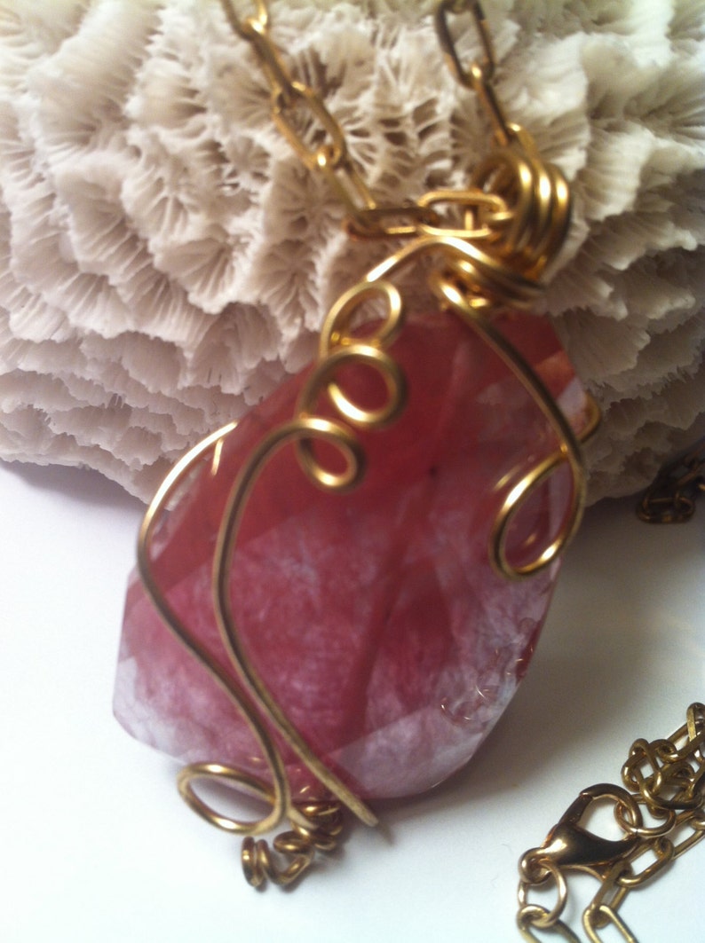 May include: A pink and white gemstone pendant wrapped in gold wire. The pendant is hanging from a gold chain.