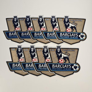 Könnte beinhalten: Eine Sammlung von Barclays Premier League Champion-Abzeichen. Jedes Abzeichen zeigt einen Löwen, einen Fußball und die Meisterschaftsjahre. Die Abzeichen sind in Gold und Blau gehalten.