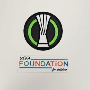 Puede incluir: Dos parches bordados. El parche superior es un círculo negro con un borde verde y un diseño blanco y negro. El parche inferior es rectangular con el texto "UEFA FOUNDATION for children" en varios colores.