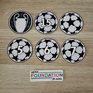 Op de afbeelding: Zes ronde patches met zwart-witte voetbalpatronen, waarvan er één een trofee-icoon bevat en andere de cijfers 15, 5, 6 en 7 tonen. Een rechthoekige patch eronder bevat de tekst UEFA FOUNDATION for children in kleurrijke letters.