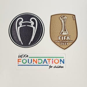 Puede incluir: Tres parches con logotipos relacionados con el fútbol. Uno es un parche circular en blanco y negro con un diseño de trofeo. Otro es un parche dorado en forma de escudo con el logotipo de la FIFA World Champions 2023. El tercero es un parche rectangular con el texto "UEFA FOUNDATION for children."
