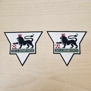 May include: Two triangular fabric patches featuring the F.A. Premier League logo. Each white patch displays a black lion wearing a red crown, resting a paw on a red and white soccer ball, with the text THE F.A. PREMIER LEAGUE in green.