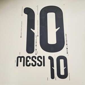 Op de afbeelding: Zwart-witte warmteoverdracht vinylvellen met het nummer "10" en de naam "MESSI" erop gedrukt. Het nummer "10" is groot en vetgedrukt, met een gestippeld patroon bovenaan. De tekst "OFFICIAL LICENSED PRODUCT" staat er ook op.