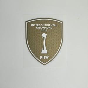 Könnte beinhalten: Ein schildförmiger Aufkleber mit dem Text "INTERCONTINENTAL CHAMPIONS 2025" und "FIFA" in Weiß. Das Schild hat einen goldfarbenen, strukturierten Hintergrund und einen weißen Rand, mit einer weißen Trophäe in der Mitte.