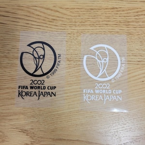 Puede incluir: Dos parches termoadhesivos en blanco y negro con el logotipo de la Copa Mundial de la FIFA 2002. El logotipo presenta una figura estilizada de una persona sosteniendo un balón de fútbol. El texto "2002 FIFA World Cup Korea Japan" está impreso debajo del logotipo. El texto "© 1999 FIFA TM" está impreso a la derecha del logotipo.
