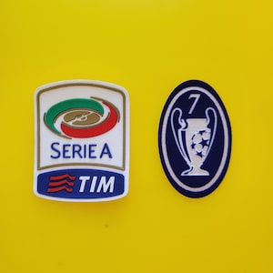 Puede incluir: Dos parches sobre fondo amarillo. Uno es un rectángulo blanco con el logo de la Serie A y el texto "SERIE A TIM". El otro es un óvalo con un trofeo y el número "7".