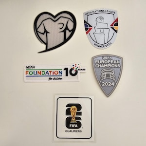 Può includere: Una collezione di toppe e adesivi a tema sportivo. Include una toppa a forma di cuore, una toppa UEFA Nations League Winner 2025, una toppa UEFA Foundation for Children 10 Years, una toppa UEFA European Champions 2024 e un adesivo FIFA Qualifiers.