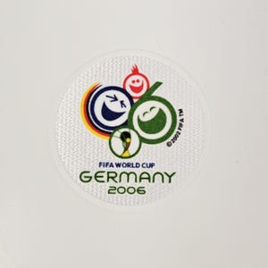 Può includere: Toppa circolare con il logo FIFA World Cup Germany 2006. Il design include facce sorridenti di vari colori, un pallone da calcio e il testo "FIFA WORLD CUP GERMANY 2006". La toppa ha un bordo bianco strutturato.