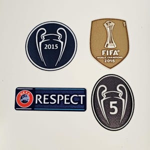Può includere: Quattro toppe con disegni sportivi. Una è un cerchio blu scuro con un contorno bianco e il numero "2015". Un'altra è uno scudo dorato con "FIFA WORLD CHAMPIONS 2015". Una toppa rettangolare recita "UEFA RESPECT". L'ultima toppa è un ovale scuro con il numero "5".