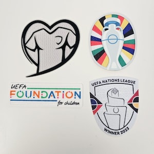 Puede incluir: Cuatro parches con diseños relacionados con el deporte. Uno tiene forma de corazón con un puño en su interior. Otro es un óvalo con un trofeo. Un parche rectangular dice "UEFA FOUNDATION for children". El último parche dice "UEFA NATIONS LEAGUE WINNER 2023."
