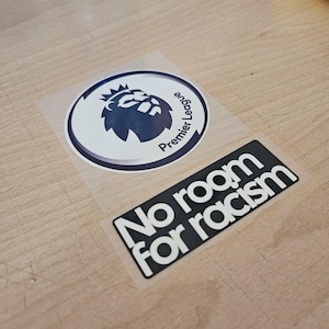 Op de afbeelding: Twee stickers op een transparante achtergrond. De ene is een rond Premier League logo met een donkerblauw silhouet van een leeuwenkop. De tweede is een rechthoekige zwarte sticker met witte tekst No room for racism in een vetgedrukt lettertype.