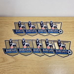 Puede incluir: Un conjunto de pegatinas de campeón de la Premier League Barclays. Cada pegatina presenta un escudo dorado con un león, un balón de fútbol y los años de campeonato. Dispuestas sobre una superficie de madera clara.