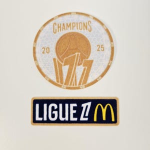 Op de afbeelding: Twee patches: een cirkelvormige patch met "CHAMPIONS" en een basketbalafbeelding, en een rechthoekige patch met "LIGUE 1" en een geel "M"-logo. Beide patches hebben een getextureerd uiterlijk.