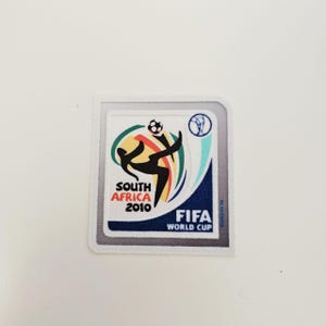 Peut inclure: Un écusson carré avec le logo de la Coupe du Monde de la FIFA 2010. Le design comprend une silhouette stylisée frappant un ballon de football, avec le texte "SOUTH AFRICA 2010" et "FIFA WORLD CUP". Le patch a une bordure blanche.