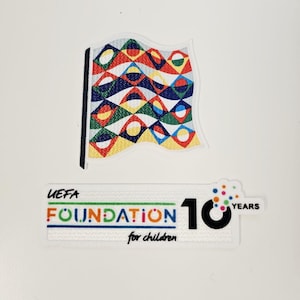 Puede incluir: Dos parches: uno con forma de bandera con un diseño geométrico colorido, y otro parche rectangular con el texto "UEFA FOUNDATION for children" y el número "10 YEARS".
