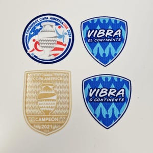 Op de afbeelding: Vier patches met voetbalthema's. Eén ronde patch met de tekst "CONMEBOL COPA AMERICA USA 2024". Twee schildvormige patches met het woord "VIBRA" en de tekst "EL CONTINENTE" of "O CONTINENTE". De vierde patch is goudkleurig en heeft de tekst "CAMPEÓN 2021".