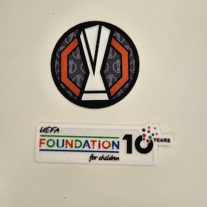 Puede incluir: Dos parches bordados. El parche superior es circular con un borde negro, un diseño blanco y naranja y un fondo gris. El parche inferior es rectangular con el texto "UEFA FOUNDATION for children" y el número "10 YEARS".