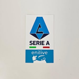 Könnte beinhalten: Ein rechteckiger Aufkleber mit dem Serie A-Logo. Das Logo ist ein blaues stilisiertes "A" über einer schwarzen und blauen Raute. Darunter steht der Text "SERIE A" mit grünen und roten Balken. Der untere Teil hat den Text "enilive" und eine weiße Grafik.