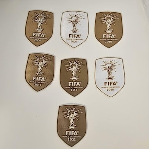 Può includere: Una collezione di toppe FIFA Campioni del Mondo. Ogni toppa, a forma di scudo, presenta il logo FIFA e l'anno della Coppa del Mondo. Le toppe sono dorate e bianche, con gli anni 2006, 2010, 2014, 2018 e 2022.