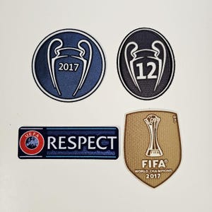 May include: Four patches with football-related logos. Two circular patches with a trophy design and the numbers "2017" and "12". A rectangular patch with "UEFA RESPECT" text. A gold shield-shaped patch with "FIFA WORLD CHAMPIONS 2017".