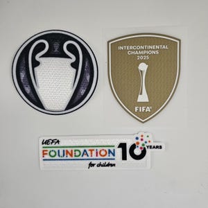 Könnte beinhalten: Drei Aufnäher: ein schwarz-weißes Champions-League-Logo, ein gold-weißes FIFA Intercontinental Champions 2025-Abzeichen und ein mehrfarbiges UEFA Foundation for Children 10 Years-Abzeichen.