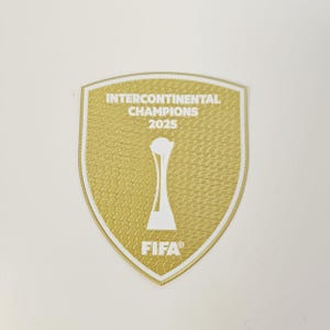 Op de afbeelding: Een goudkleurige patch in schildvorm, met de tekst "INTERCONTINENTAL CHAMPIONS 2025" en "FIFA" in het wit. Een witte trofee-afbeelding staat in het midden. De patch heeft een getextureerd oppervlak.