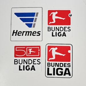 Può includere: Quattro adesivi quadrati. Uno presenta la parola "Hermes" sotto un design ad ala blu e grigio. Tre adesivi hanno le parole "BUNDES LIGA" e una grafica di giocatore di calcio, due in rosso e bianco, uno con "50" in rosso.