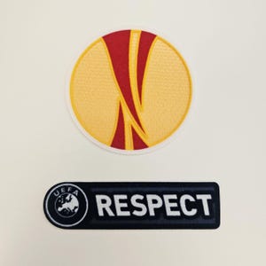 Könnte beinhalten: Zwei gestickte Aufnäher. Der obere Aufnäher ist ein gelber Kreis mit einem roten und gelben Design. Der untere Aufnäher ist ein schwarzes Rechteck mit dem Wort "RESPECT" in Weiß und einem UEFA-Logo.