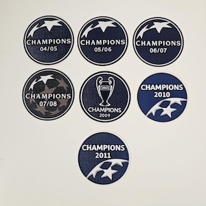 Könnte beinhalten: Eine Sammlung runder Aufnäher mit dem Champions League-Logo und dem Wort "CHAMPIONS" sowie den Jahreszahlen 04/05, 05/06, 06/07, 07/08, 2009, 2010 und 2011. Die Aufnäher sind hauptsächlich marineblau und weiß.