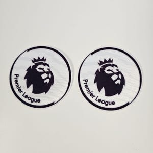 Op de afbeelding: Twee ronde patches met het Premier League-logo. Het logo toont een zwarte leeuw met een kroon en de woorden "Premier League" in zwarte tekst rond de witte cirkel.