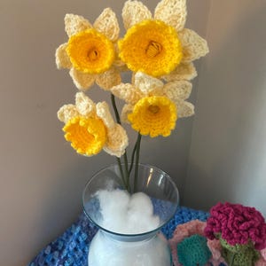 May include: A bouquet of crocheted daffodils in a clear glass vase with white stuffing. The daffodils are yellow and white and have a realistic appearance.