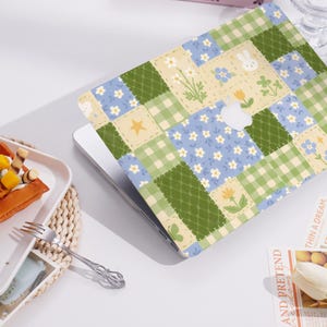 May include: A laptop computer with a colorful patchwork design cover. The design features a variety of floral patterns, gingham checks, and a small bunny rabbit. The laptop is on a white surface.