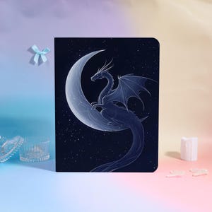 May include: A dark blue notebook featuring a detailed illustration of a dragon perched on a crescent moon against a starry night sky. The dragon is depicted in shades of blue and white, with intricate wing and scale details.