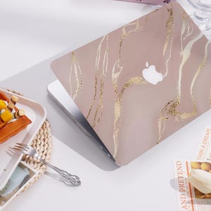 May include: A laptop with a blush pink cover featuring a gold marble design. The Apple logo is visible on the cover. The laptop is open, revealing the silver keyboard and screen.