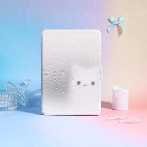 Pode incluir: Uma capa retangular branca com um design de rosto de gato, com um nariz pequeno e dois pontos para os olhos. A capa tem uma aparência molhada com gotas de água. Uma fita azul clara e uma pequena vela branca também estão na imagem.