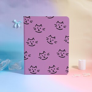 Puede incluir: Una funda de tableta lavanda con un patrón repetido de caras de gato sonrientes negras. La funda es rectangular y está en posición vertical. Un pequeño lazo azul es visible en el fondo, junto con una pequeña vela y cristal decorativo.