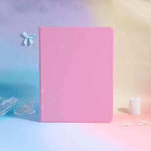 May include: A solid pink tablet case stands upright against a gradient background of blue, purple, and yellow. A small blue bow is attached to the background. Decorative glass bowls and a white candle are in the foreground.