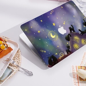 May include: A laptop skin featuring a dark blue night sky with yellow stars, a crescent moon, and several black cat silhouettes perched on a rooftop. The design includes a white Apple logo cutout in the center and glowing yellow windows on the house.