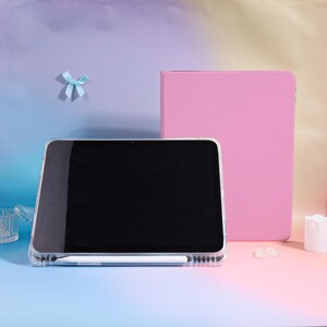 Pode incluir: Uma capa de tablet transparente com uma caneta branca e uma capa de tablet rosa. A tela do tablet &eacute; preta. Uma pequena fita azul est&aacute; no fundo. O fundo &eacute; um gradiente de azul, rosa e amarelo.
