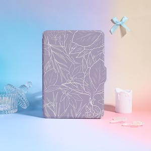 May include: A lavender-colored case with a white floral design. The case has a magnetic closure and is rectangular. A light blue bow is tied at the top. A small white candle and a glass container are also in the image.