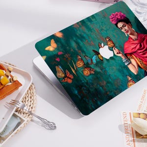 May include: Laptop skin featuring a portrait of a woman with a floral headpiece and red shawl, surrounded by butterflies. The background is a textured teal and green. The Apple logo is integrated into the design. The overall theme is art and design.